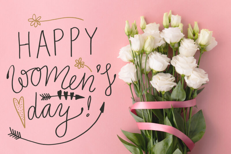 From above Happy Women's day words and bunch of flowers with ribbon on pink background.